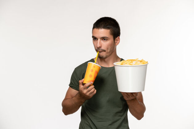 年轻男性正面图身穿绿色t恤的年轻男性手持土豆cips 在白墙上喝苏打水电影电影院男性孤独电影成人酒精汽水