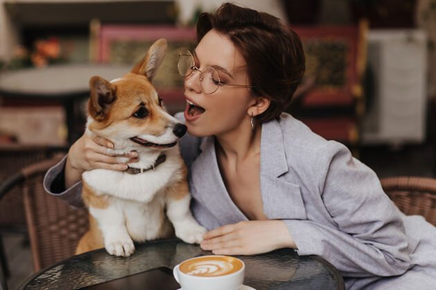 女性戴着眼镜的迷人女人在街上的咖啡馆休息时和她的狗玩穿着灰色夹克的迷人女人亲吻高尔基犬短发外部时尚