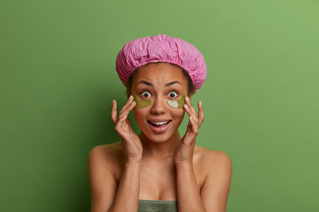 面部令人惊讶的黑皮肤女性的肖像触摸柔软的皮肤 应用眼罩减少浮肿和细纹 戴浴帽 身体周围柔软的毛巾 隔离在绿色的墙壁上浴缸触摸惊喜