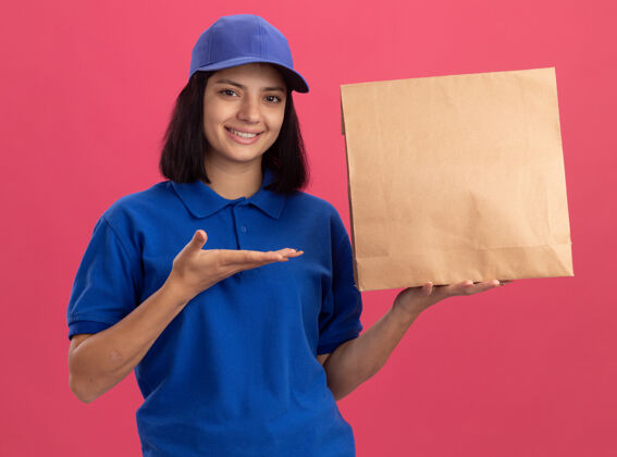 送货身穿蓝色制服 头戴礼帽的年轻送货女孩展示纸包装 手臂微笑自信地站在粉色的墙上年轻帽子手臂
