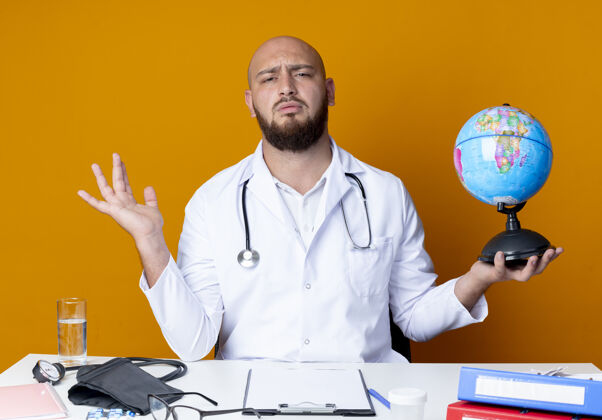 医疗困惑的年轻秃头男医生穿着医用长袍和听诊器坐在办公桌旁 手持医疗工具 手持地球仪 将手分开放在橙色背景上蔓延秃头地球仪