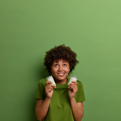 乳制品高兴的深色皮肤的女人拿着一罐罐的有机天然酸奶 准备早餐的原料 浓缩在上面 戴着帽子 露出牙齿般的微笑 穿着绿色t恤人与健康的营养理念乐观青少年专注