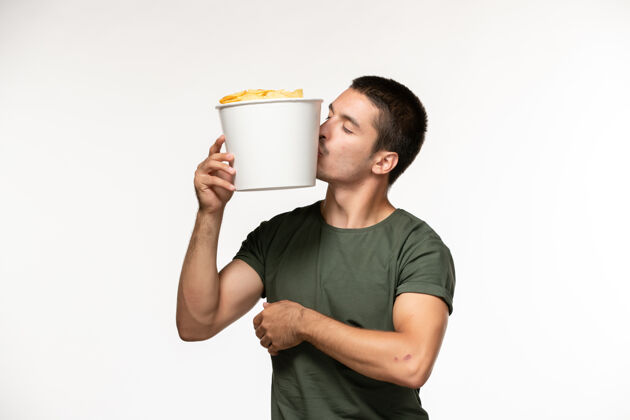 肖像正面图身穿绿色t恤的年轻男性手持土豆cips站在白色的墙上孤独的人土豆杯子视图