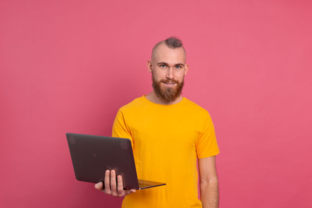 笔记本一个微笑的男人在粉色背景下用笔记本电脑工作使用电脑休闲黄色