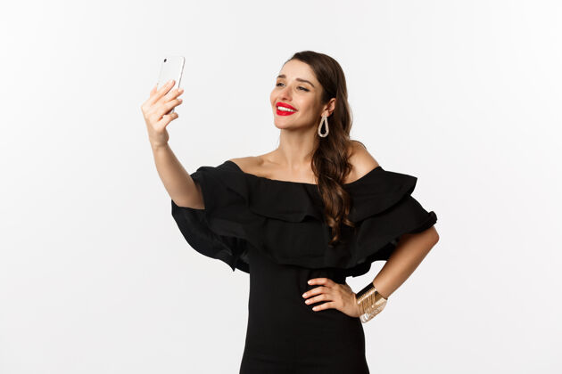 美丽身着黑色礼服的美女在派对上自拍 拿着智能手机站在白色背景上年轻视频通话优雅