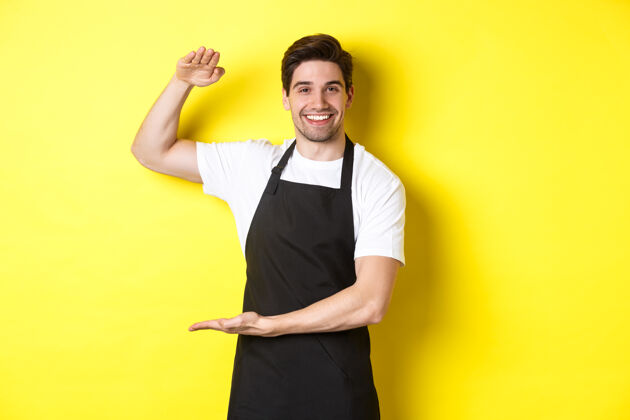 表情面带微笑的英俊的咖啡师 站在黄色的背景上 展示一些长的或大的东西尺码围裙男士