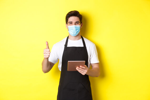 模特covid-19的概念 小型企业和流行病友好的服务员戴着医用面罩和黑色围裙 竖起大拇指 用黄色背景的数字平板电脑接受订单男士外科面膜站立