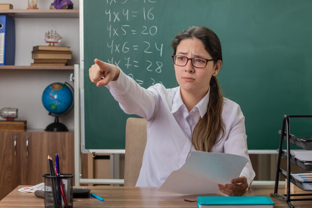房子戴眼镜的年轻女教师坐在课桌旁 用食指指着教室黑板前的作业黑板前面页