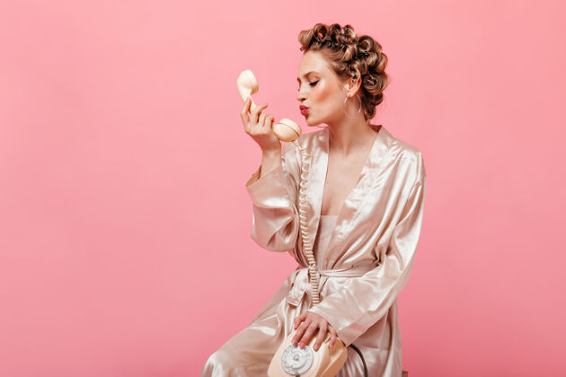 肖像穿着丝绸家装的卷发女人坐在粉色墙上的椅子上亲吻手机金发时尚家庭主妇