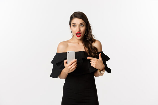 口红网上购物概念穿着黑色连衣裙 化妆 手指着手机 站在白色背景下的时尚女性女人美丽服装
