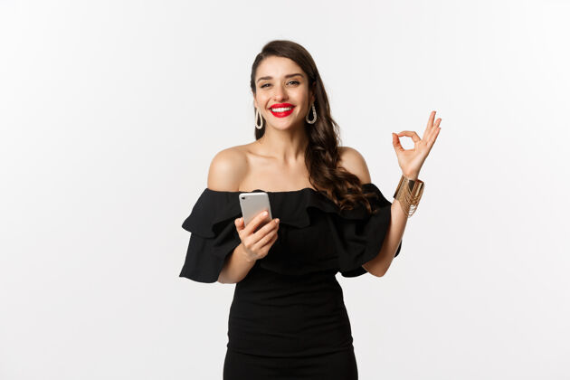 女人网上购物概念穿着时髦的黑色连衣裙 化妆 显示同意登录并使用手机应用程序 白色背景的女人表情年轻美丽