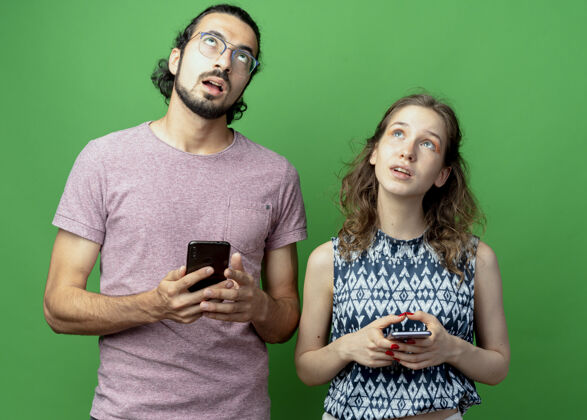 拼图年轻的一对男女 举着智能手机站在绿色的墙上 疑惑地抬头望去站着而女人