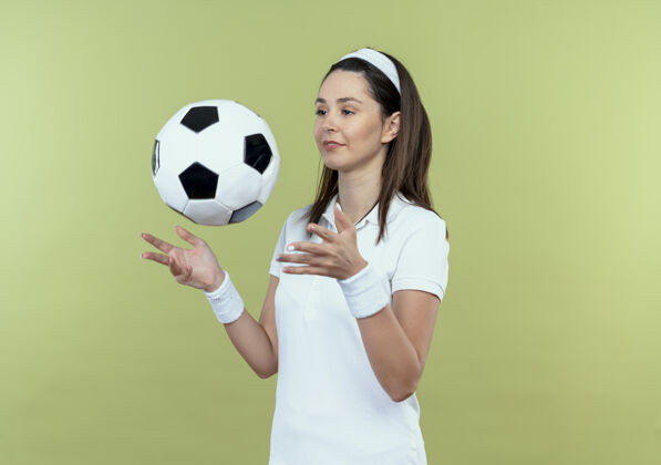 健身戴着头巾的年轻健身女士 微笑着 自信地站在轻盈的墙壁上 扔着足球球头带足球