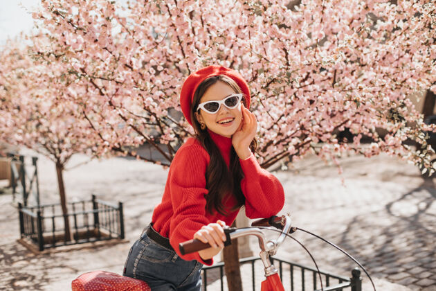 花乐观的女孩穿着红色毛衣 戴着眼镜 斜靠在樱花背景下的自行车上穿着时尚贝雷帽的迷人女人在花园里微笑华丽天温柔