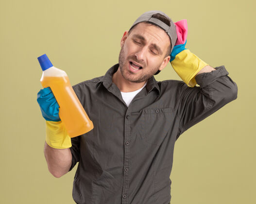 穿年轻的保洁员穿着休闲服 戴着橡胶手套 手里拿着一瓶清洁用品和抹布 站在绿色的墙边 看上去很困惑 很焦虑焦虑瓶子衣服