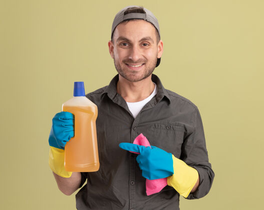 积极年轻的清洁工穿着休闲服 戴着橡胶手套 戴着帽子 手里拿着装有清洁用品和抹布的瓶子 站在绿色的墙边 脸上洋溢着快乐和积极的微笑男人衣服穿