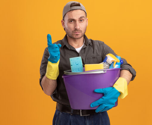 男人年轻人穿着休闲服 戴着橡胶手套 戴着帽子 手里拿着水桶 手里拿着清洁工具 表情自信 食指站在橙色的墙上表情水桶帽子