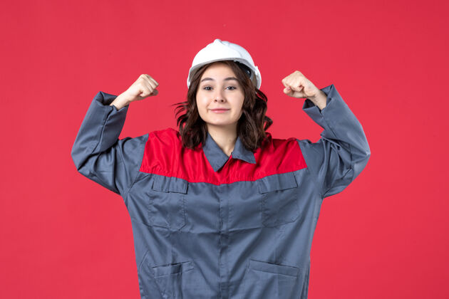 前面前视图自信微笑的女建筑工人制服与安全帽 并显示她的肌肉上孤立的红墙成人女建设者吸引力