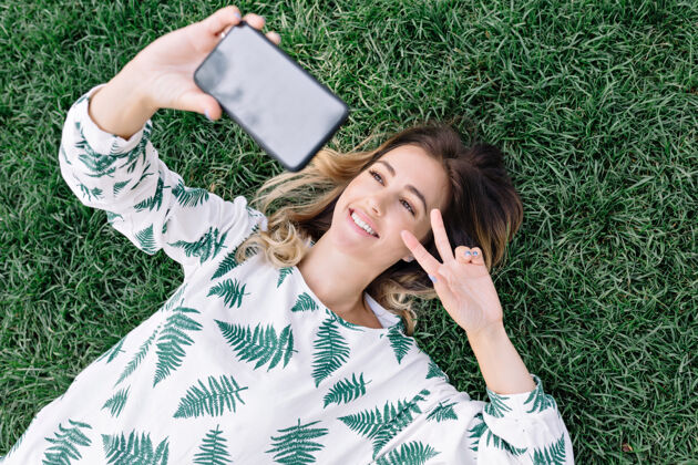 卷曲美丽的女人躺在公园的草地上 用手机自拍 并显示和平的迹象休闲夹克年轻