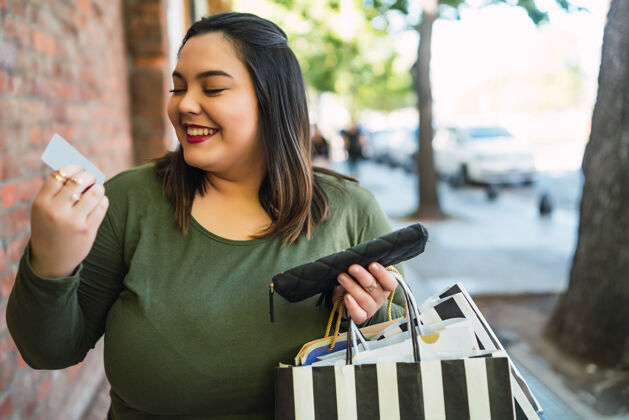 曲线街上手持信用卡和购物袋的大号年轻女子的画像购物和销售概念微笑包城市