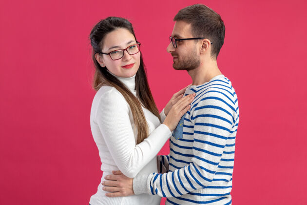 年轻年轻漂亮的情侣幸福的男人和女人穿着休闲服戴着眼镜幸福的恋爱在一起拥抱庆祝情人节的概念站在粉红色的墙上男人穿着庆祝