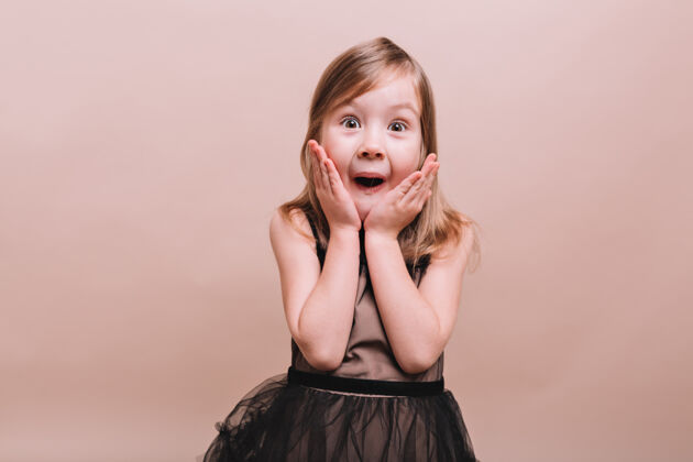 孩子伟大可爱的小女孩在米色墙上摆出奇妙的惊奇的表情小女孩张开鼠标摆出姿势 手放在脸上 真实的情绪 文字的地方快乐童年美丽