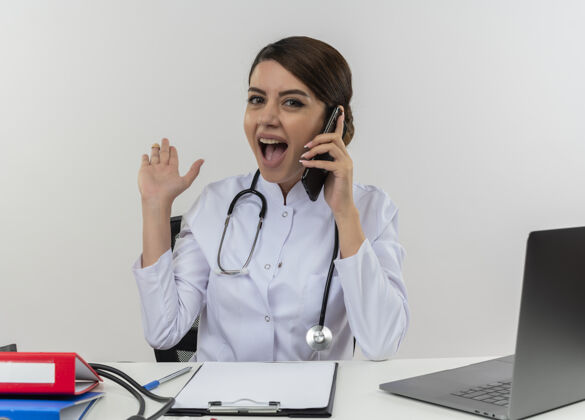 穿着令人印象深刻的年轻女医生穿着医用长袍和听诊器坐在办公桌旁 拿着医疗工具和笔记本电脑在讲电话 显示空手孤立在白墙上女性印象谈话