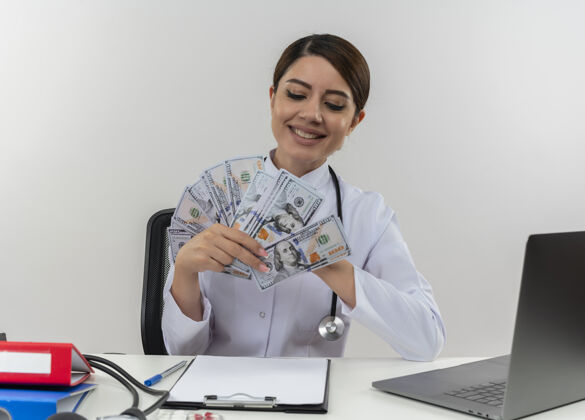 女性面带微笑的年轻女医生穿着医用长袍和听诊器坐在办公桌旁 手里拿着医疗工具和笔记本电脑 看着隔离在白墙上的钱听诊器笔记本电脑办公桌