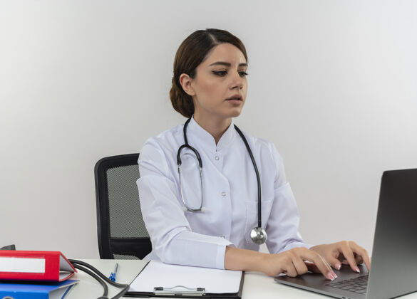 听诊器专注的年轻女医生穿着医用长袍和听诊器坐在办公桌旁 用笔记本电脑隔离在白色的墙上坐着医疗专心