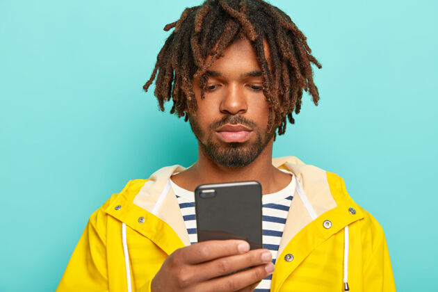 通信严肃的家伙用现代手机 上网 网上支付 穿条纹套头衫和黄色防水雨衣非洲裔衣服手机