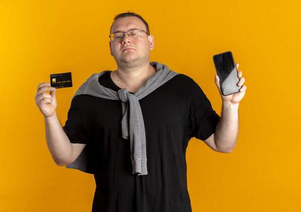 看戴眼镜的超重男子身穿黑色t恤 手持智能手机 信用卡看起来比橙色更自信卡片信用卡黑色