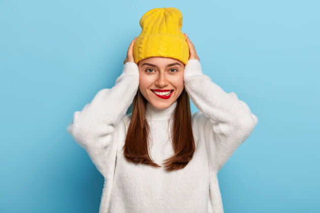 黑发快乐的女人化妆 鲜红的唇膏 双手放在头上 露出白色的牙齿 戴帽子和休闲毛衣衣服快乐穿着