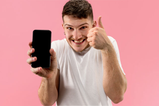 惊人一个年轻帅哥带着一张惊讶的脸在粉色屏幕上展示智能手机科技男人手机