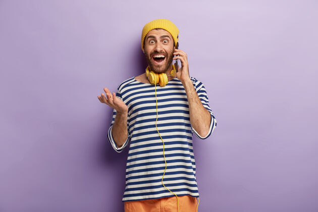 年轻好笑的嬉皮士把手机放在耳边 进行有趣的电话交谈 戴着黄色帽子和条纹套头衫通信人享受