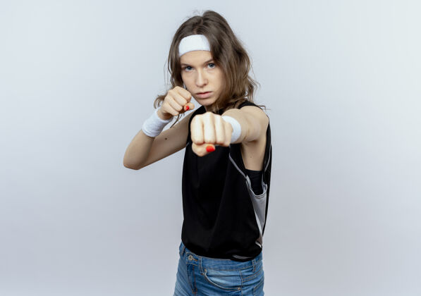 拳击手身穿黑色运动服 头戴头巾 严肃表情的年轻健身女孩站在白墙上 像个握紧拳头的拳击手握紧拳头年轻