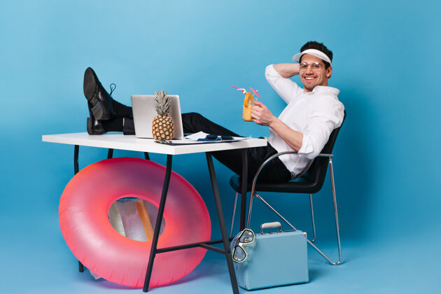 员工戴帽子的男人在度假时工作 喝鸡尾酒那个家伙正坐在桌子旁 手里拿着手提箱 充气圈 笔记本电脑和菠萝行政办公桌潜水面具