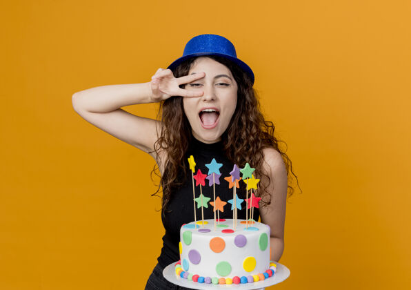 兴奋一位年轻漂亮的卷发女士 戴着节日礼帽 手里拿着生日蛋糕 高兴而兴奋地站在橙色的墙上展示着v形标志站着抱着年轻