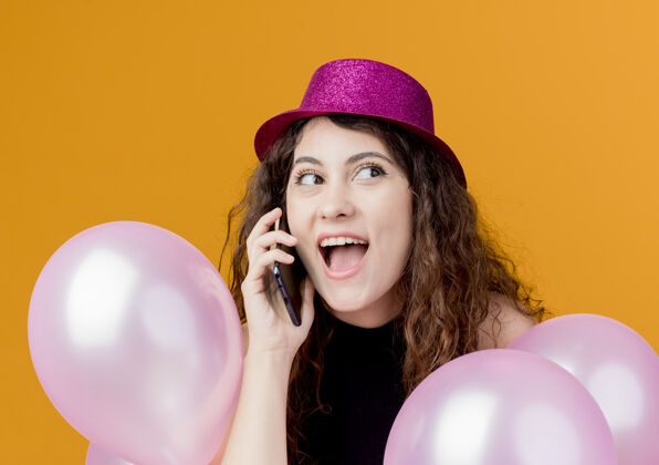 空气一个年轻漂亮的女人 卷发 戴着节日帽子 手里拿着一堆气球 用手机聊天 站在橘色的墙上 快乐而兴奋的生日聚会站着卷发一群