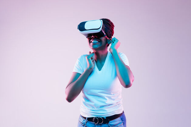 霓虹灯年轻的非洲裔美国妇女的肖像玩在虚拟现实眼镜在霓虹灯梯度背景耳机多彩表情
