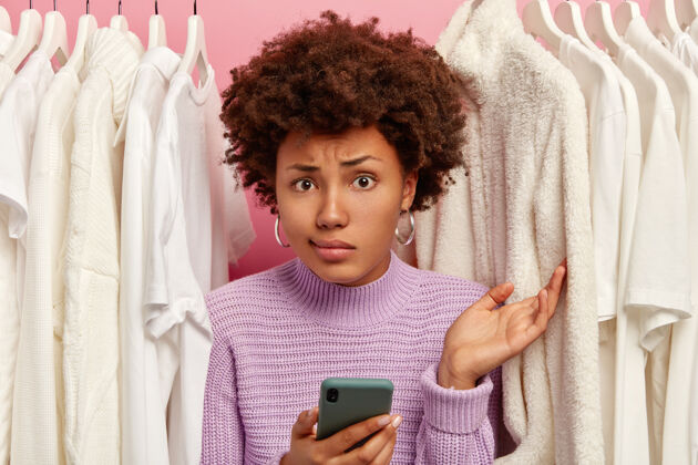 手机犹豫不决的糊涂女人用质疑的表情举起手掌 拿着现代手机 上网购物 在挂在衣架上的白衣服中间摆姿势犹豫顾客美国