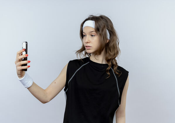 手机身穿黑色运动服 头戴头巾的年轻健身女孩站在白墙上 带着怀疑的表情看着智能手机屏幕表情年轻运动装