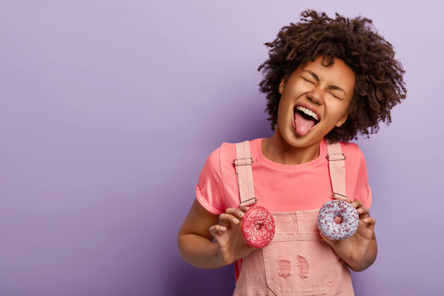 女人甜蜜的痴迷有趣的卷发女人伸出舌头 拿着两个美味的甜甜圈 穿着粉红色的衣服 隔着紫色的工作室墙黑发快乐吃