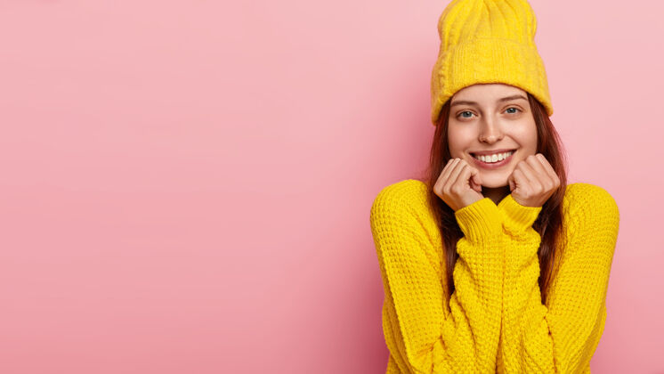高兴漂亮的女模特双手托着下巴 对着镜头温柔地微笑 戴着时髦的黄色帽子和毛衣 模特们站在粉色的墙上毛衣女孩自由空间