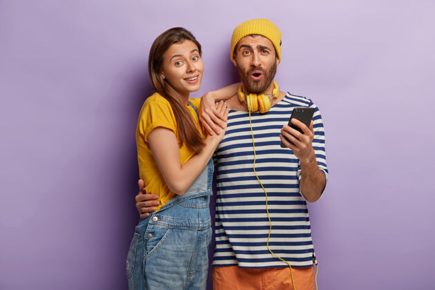 男人开朗的男女拥抱并使用现代智能手机 穿着时尚的服装 隔离在紫色的墙上聊天年轻紫色