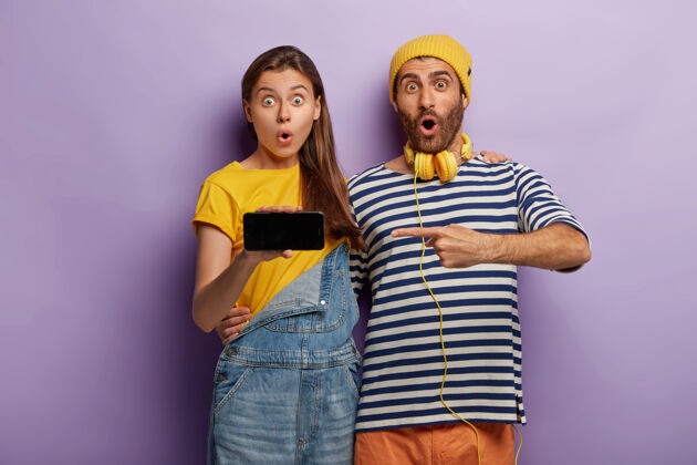 情侣兴奋惊讶的潮人指着现代的智能手机屏幕 为你的宣传内容展示模型空间 呆呆地拥抱和凝视 隔着紫色的墙壁科技广告指示灯高兴帽子