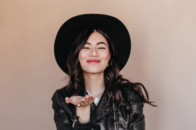 人物幸福的亚洲女人戴着帽子在米色背景上做手势浪漫卷曲的日本女人穿着皮夹克的摄影棚拍摄美丽黑发表情