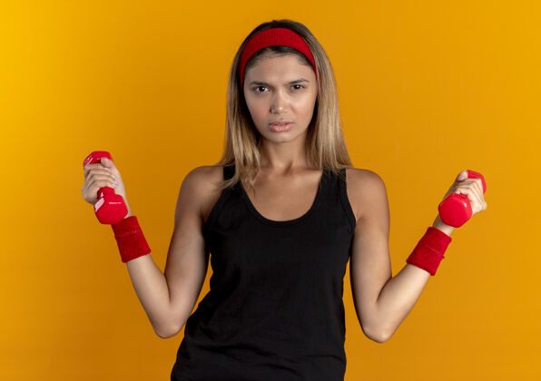 女孩身穿黑色运动服 头戴红色头巾的年轻健身女孩 正脸举着哑铃站在橙色的墙上锻炼身体头带工作年轻