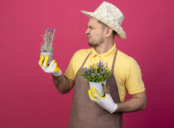 抱着年轻的园丁 穿着连体衣 戴着帽子 戴着工作手套 手里拿着盆栽植物 看着它们感到困惑和不快粉色花园植物