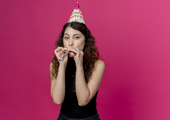 女人年轻漂亮的女人卷发在节日帽子吹哨子快乐和积极的生日派对概念站在粉红色的墙上吹帽子积极