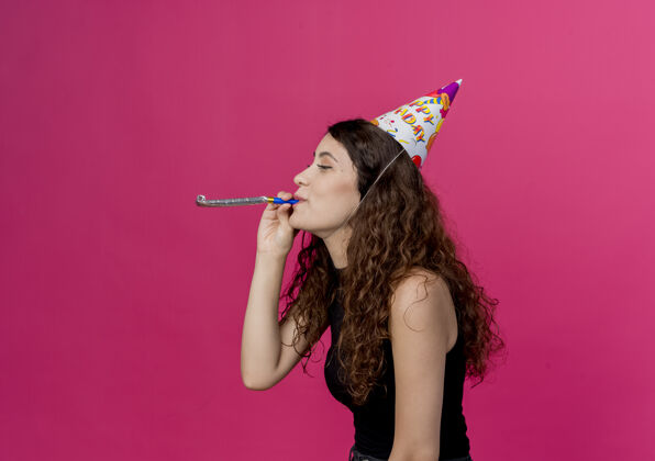 站年轻漂亮的女人卷发在节日帽子吹口哨生日聚会的概念站在粉红色的墙上哨子帽子卷发